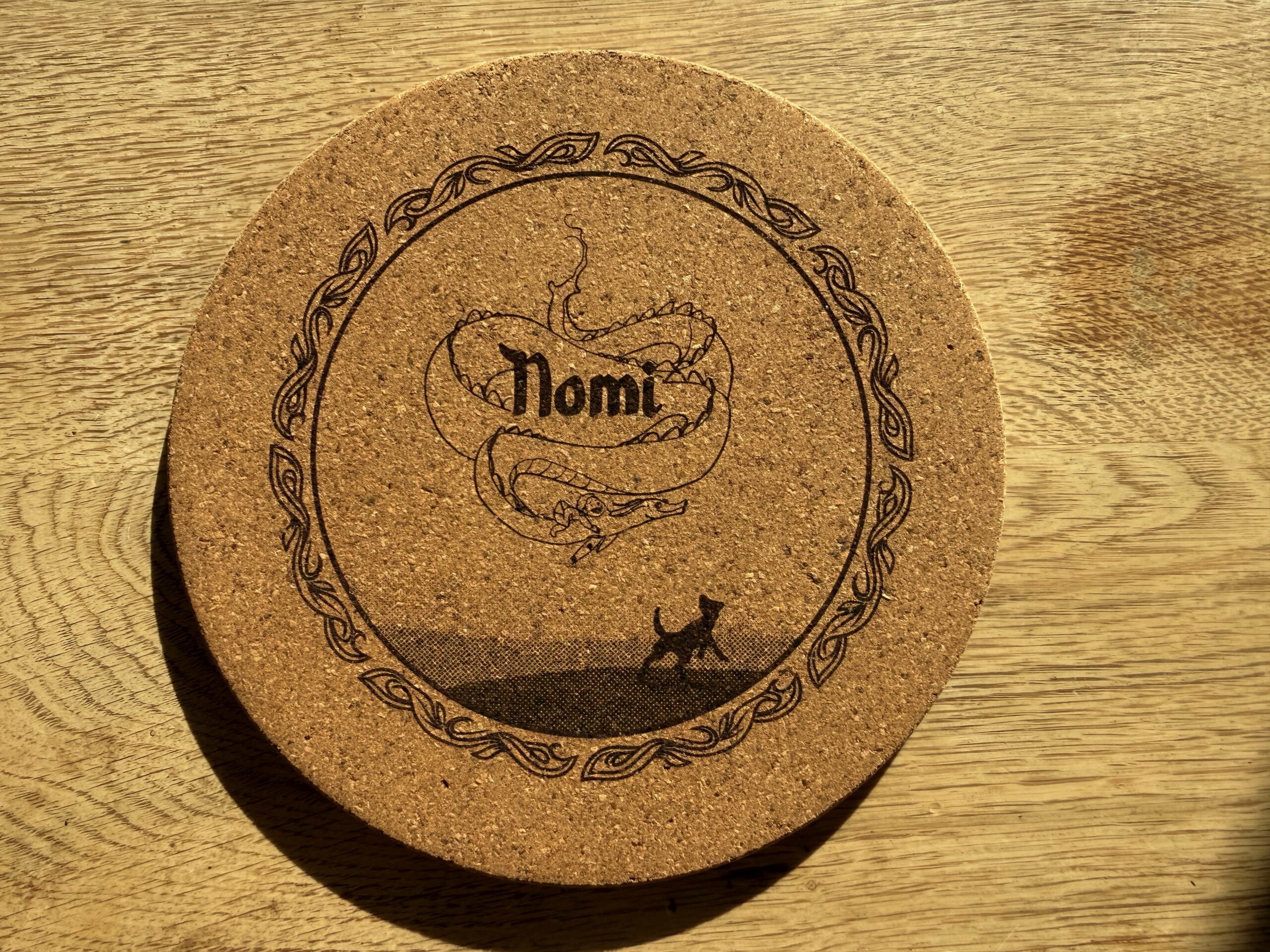 Cork coaster engraved with a dragon, a dog and a child's name inside a decorative border