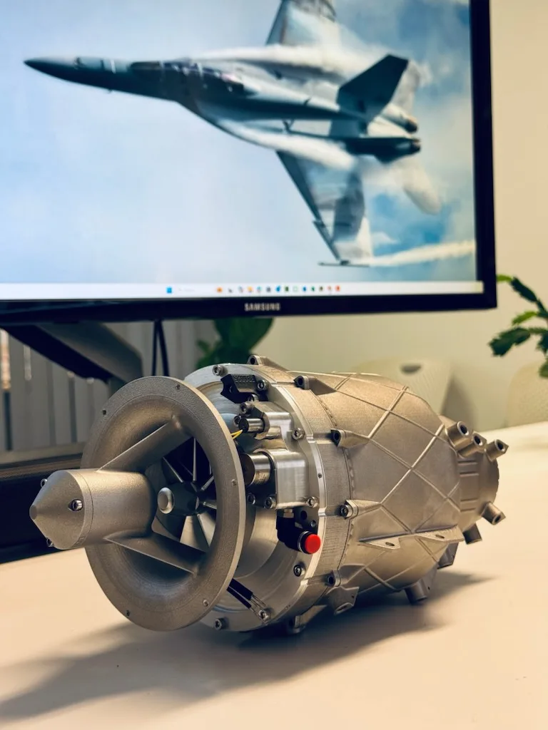 A metal Aurora Labs turbojet engine on a desk in front of a monitor showing a fighter jet