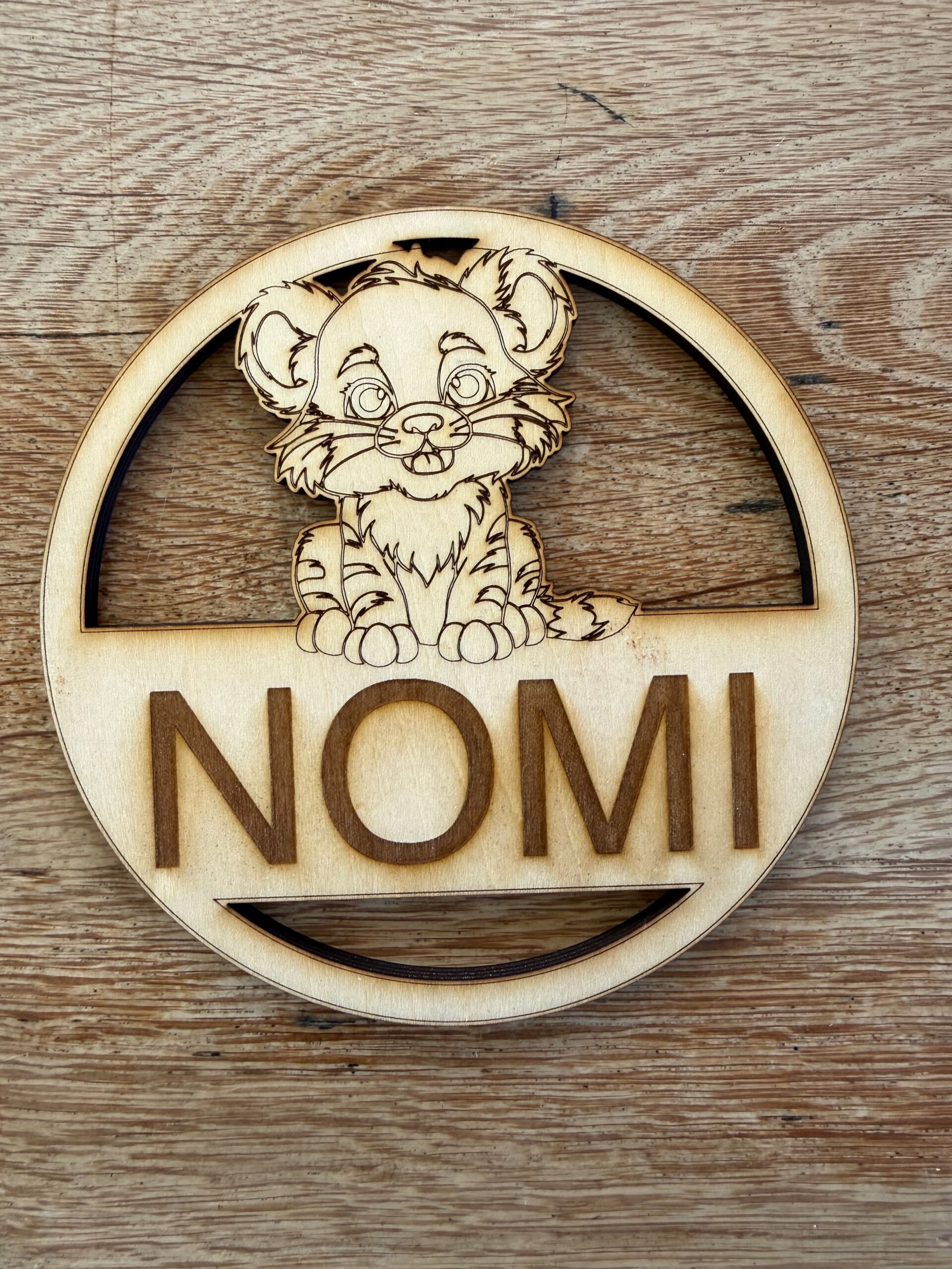 A round plywood name plate with a tiger cub engraved above a child's name