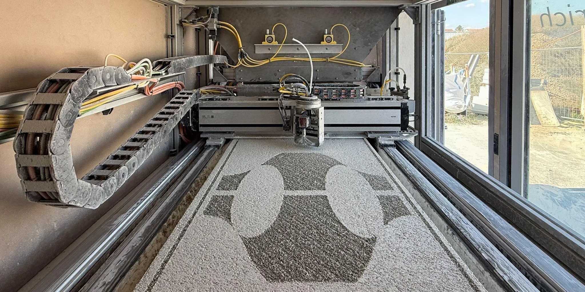 A large binder-jetting machine with a dark geometric pattern bound into a bed of pale mineral powder
