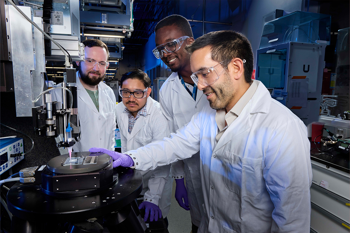 Four LLNL researchers in lab coats around a direct-ink writing printer