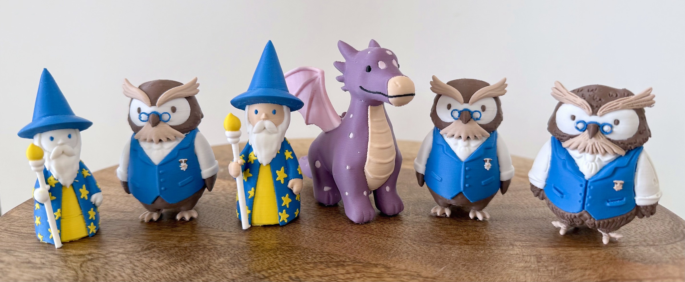 Everything the review printed, in one row: the first wizard (65 mm, white where the skin tone should have been), an owl from the Nano Banana redraw, the reprinted wizard (75 mm), the dragon (75 mm), the second redraw owl and owl A from Sonja's own drawings (60 mm). Snapmaker U1, Polymaker Panchroma Matte PLA, files from Meshy Multicolor 2.0.