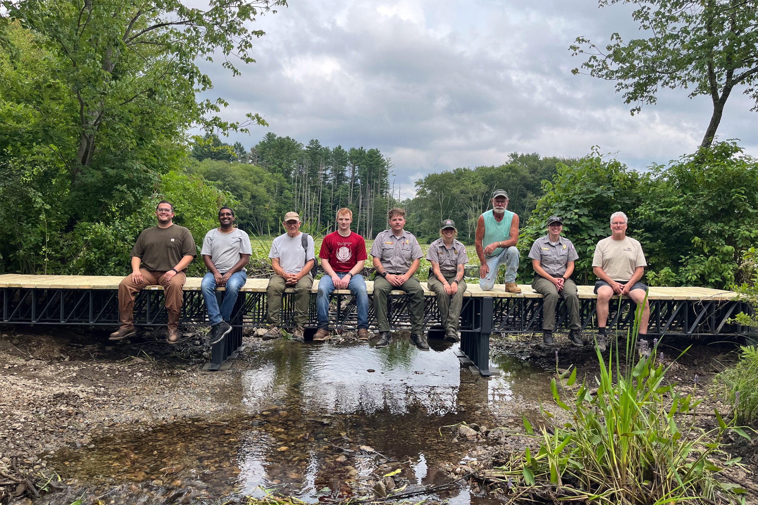 Nine people sitting on a 3D-printed recycled-composite bridge over a wetland stream