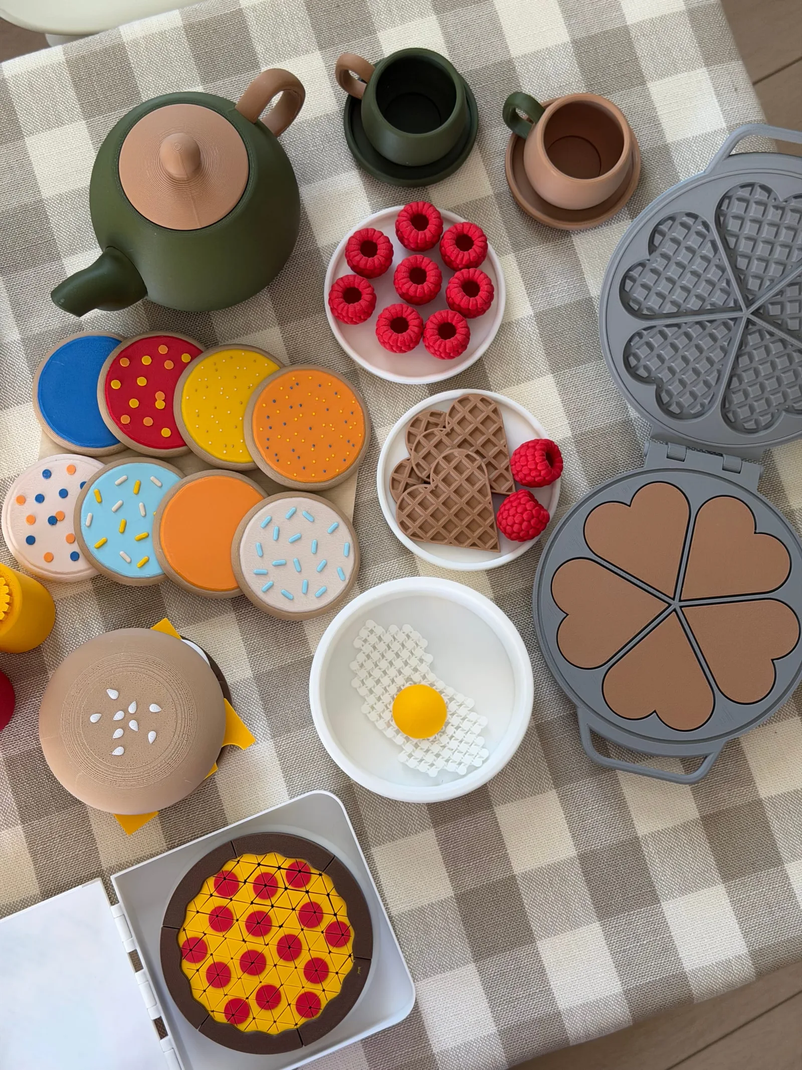 Toy teapot, cups, decorated cookies, waffles, raspberries, an egg, a burger and a pizza on a checked tablecloth