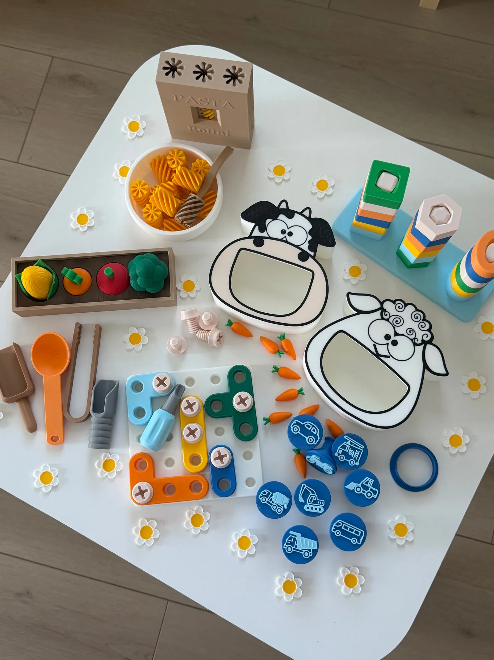 Toys spread on a white table including a pasta box and bowl, cow and sheep shapes, stacking towers and a screw board