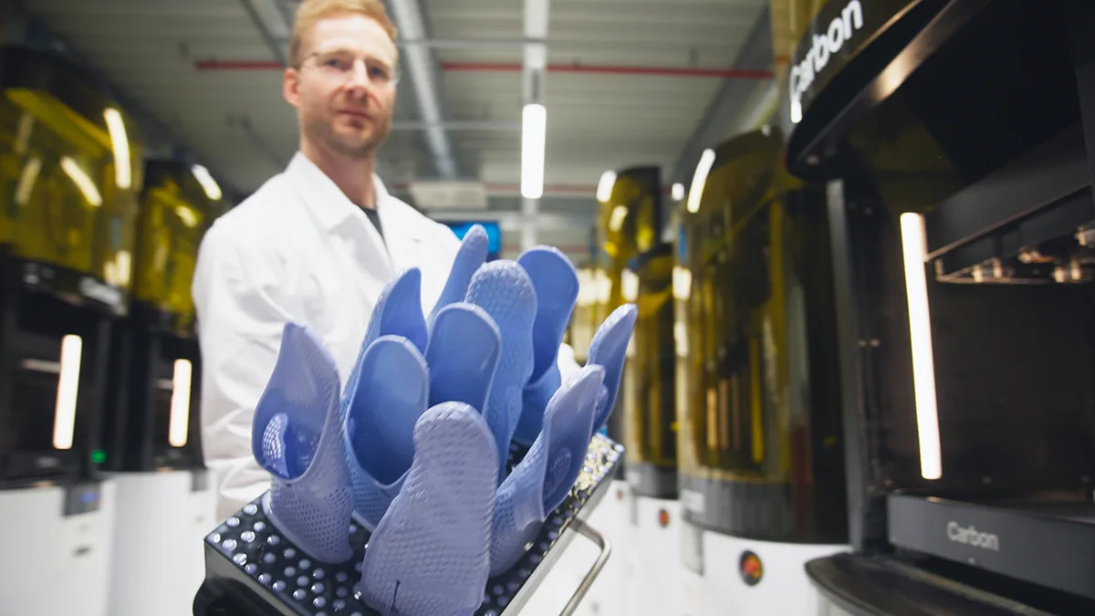 A technician in a white coat holds a tray of blue printed lattice parts beside a row of Carbon printers