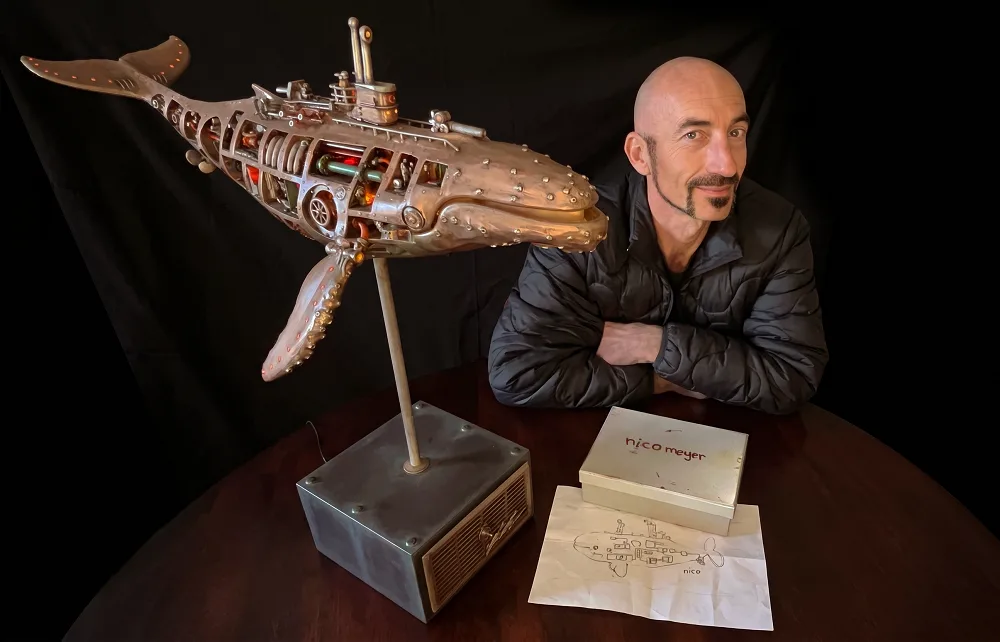 Nico Meyer sitting at a round wooden table beside his whale sculpture on a metal stand, with a box marked nico meyer and a pencil sketch of the whale in front of him