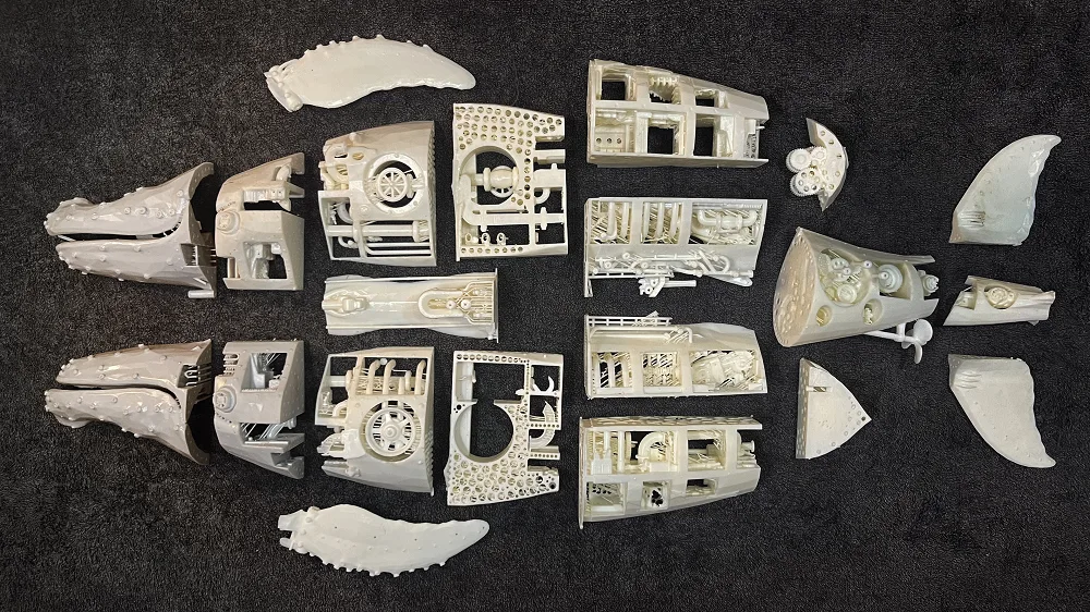 Unpainted white resin 3D printed parts of the whale sculpture laid out on a dark carpet in the shape of the whale, including head halves, detailed body sections, fins and tail flukes
