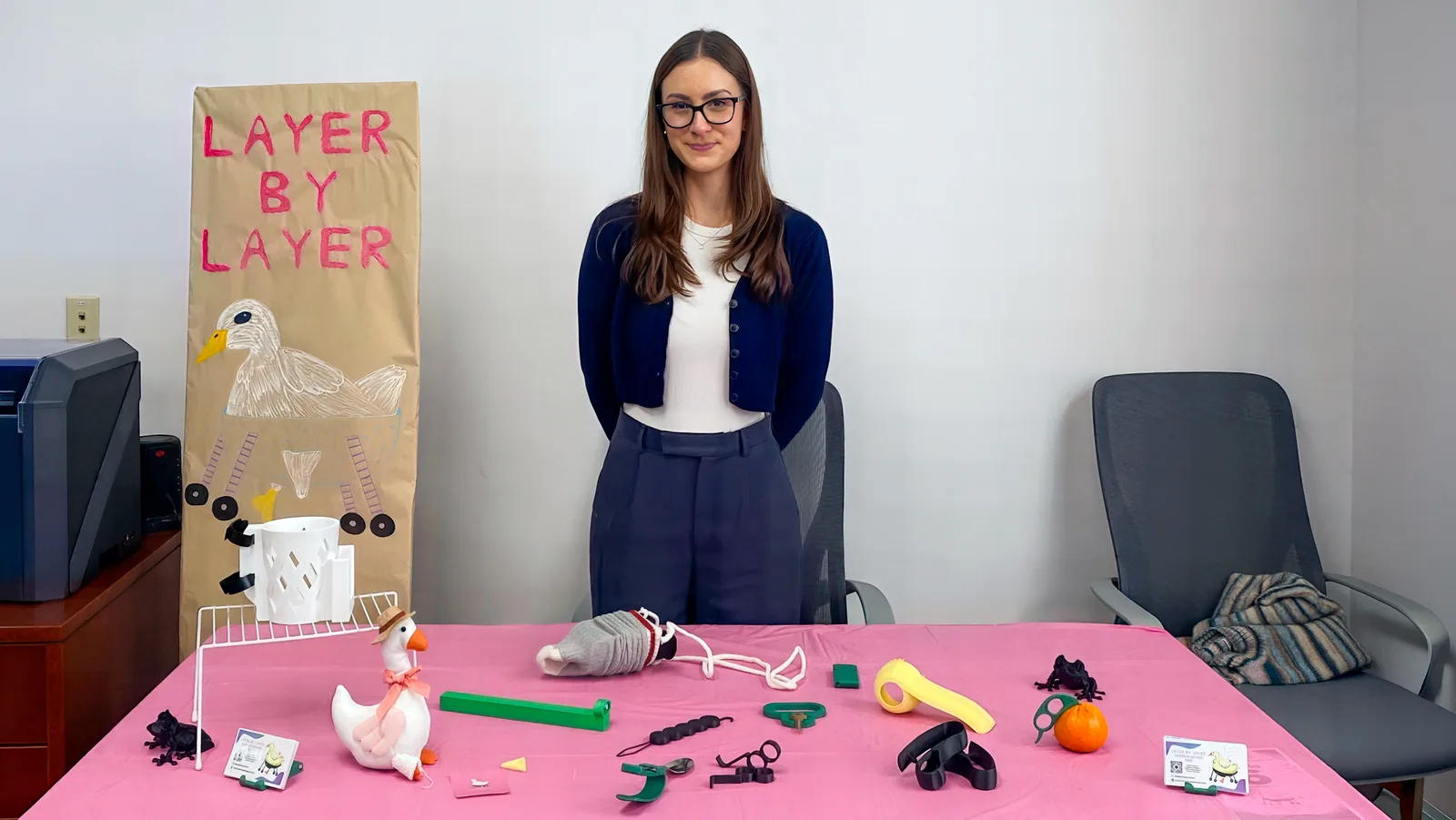Vianna Aiello standing behind a table of 3D printed assistive devices next to a Layer By Layer club banner